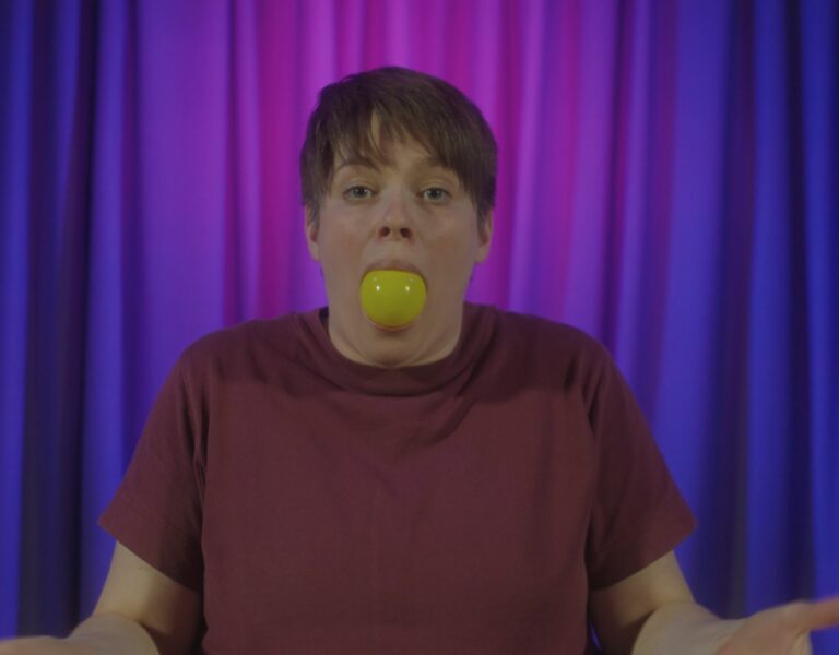 A short-haired woman with a plastic ball in her mouth, standing in front of a curtain.