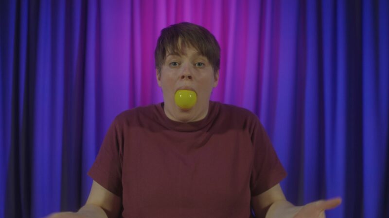 A short-haired woman with a plastic ball in her mouth, standing in front of a curtain.