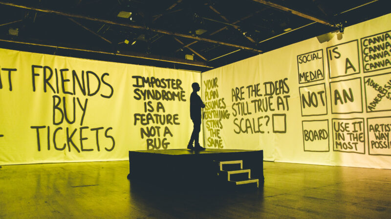 Elijah standing on a small stage in silhouette, surrounded by yellow screens.