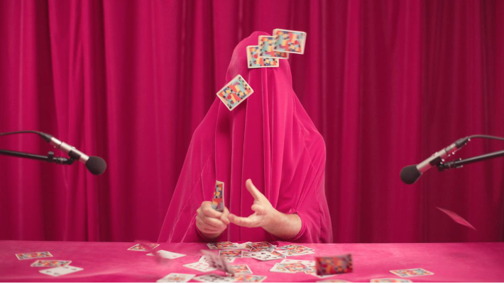 A person is draped in pink material covering themselves, apart from their hands and forearms. They sit at a table covered in a pink cloth upon which playing cards are scattered. They look as if they are attempting card tricks. Two microphones sit either side of the desk.