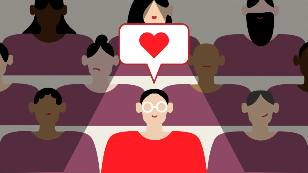 An illustration of a diverse group of people in minimalist style. One person in a red shirt is shown in the centre, smiling and wearing glasses, with a speech bubble containing a red heart icon hovering above their head. This person seems to be the focus or recipient of affection or appreciation. Surrounding this central figure are several other people with varying skin tones, hairstyles, and facial expressions, all wearing purple shirts. The group appears to be represented in a flat, simplified design. The overall composition and the heart icon suggest themes of community, connection, appreciation, or expressing positivity towards someone within a group setting.