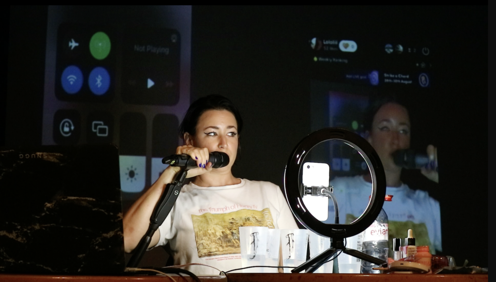 A person speaks into a microphone in a studio setting. They are illuminated by a donut light which sits in front of them.