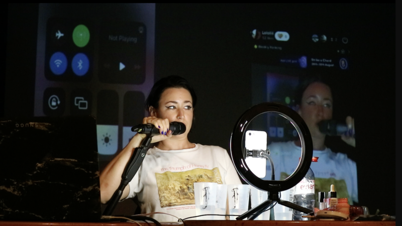 A person speaks into a microphone in a studio setting. They are illuminated by a donut light which sits in front of them.