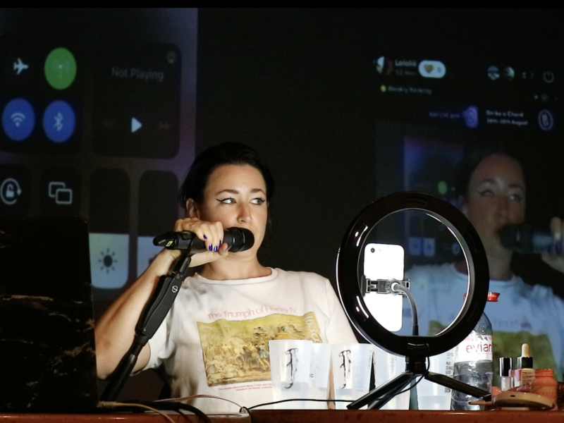 A person speaks into a microphone in a studio setting. They are illuminated by a donut light which sits in front of them.