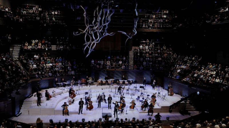 A large auditorium with the stage situated in the round. Members of Aurora Orchestra stand on stage playing their instruments, surrounded by a seated audience.