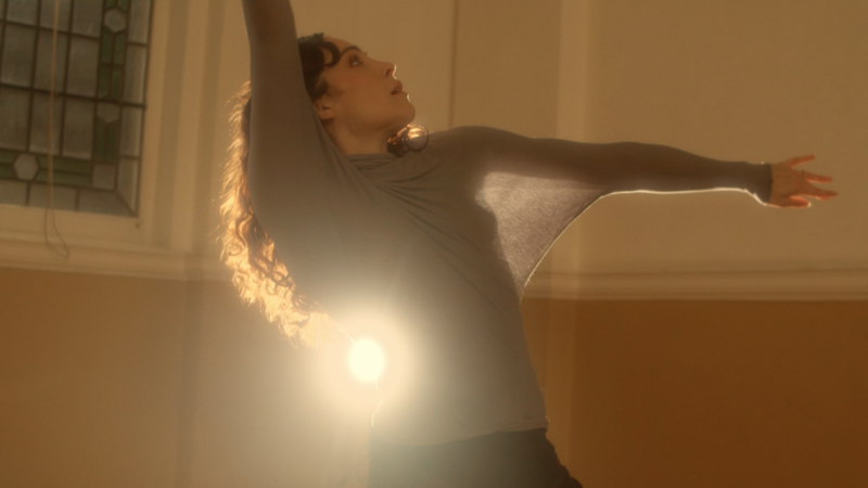 Photo of a dancer with her arms outstretched and a chink of light shining at her lower back.