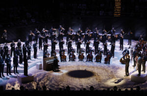 Members of a choir or orchestra stand on a stage, arranged in a half-circle.