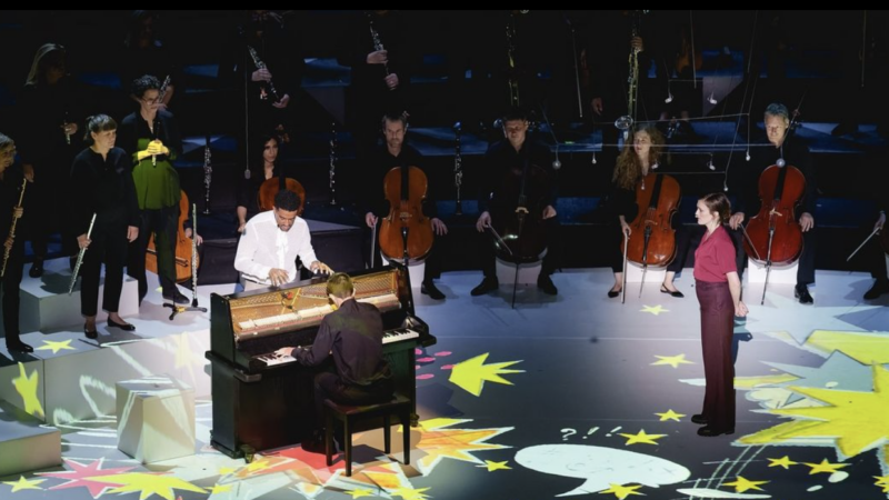A large indoor performance area with stars projected onto the floor area. A person in a white suit stands playing an upright piano. people stand around the edge of the performance area.