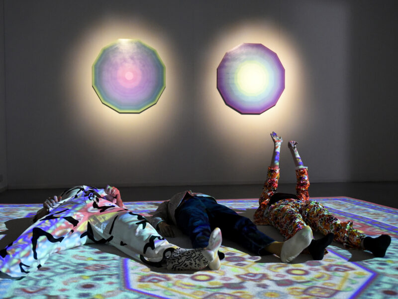 Photo of three people lying on a light projection of a colourful Islamic rug.