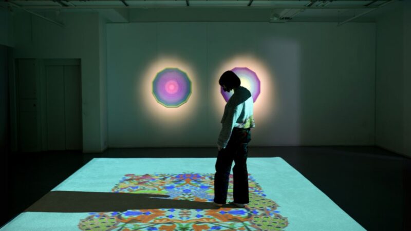 Photo of a person in silhouette standing on a light projection of a colourful Islamic rug.