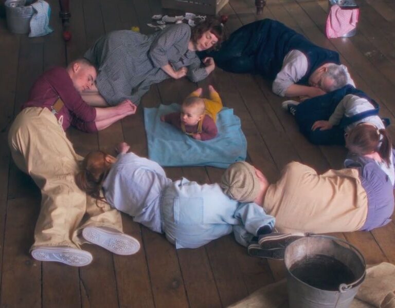 Dancers sleeping in a circle, on a wooden floor, with a baby in the centre of the circle.
