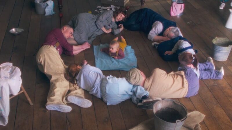 Dancers sleeping in a circle, on a wooden floor, with a baby in the centre of the circle.