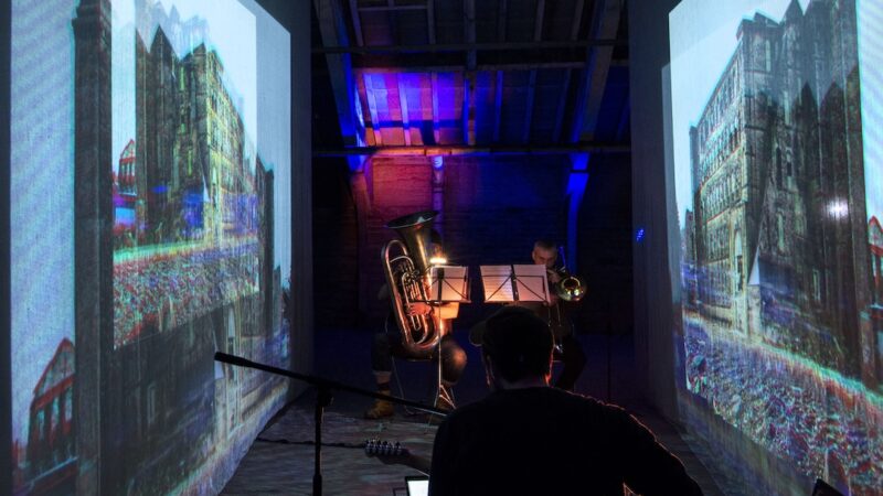 People playing brass instruments between 2 screens of of archival footage of textile mills.