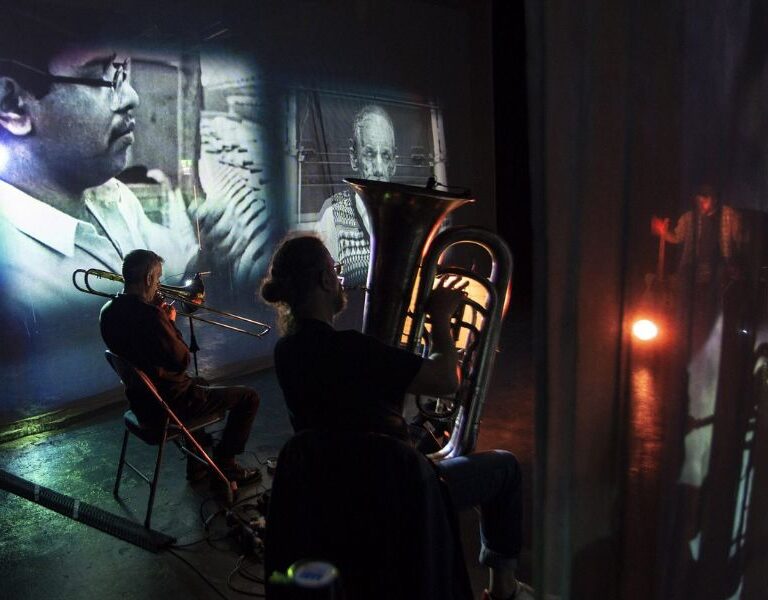 Image from the installation of 2 men playing brass instruments in front of black and white archival film footage.