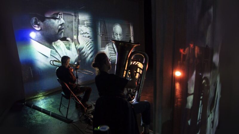 Image from the installation of 2 men playing brass instruments in front of black and white archival film footage.