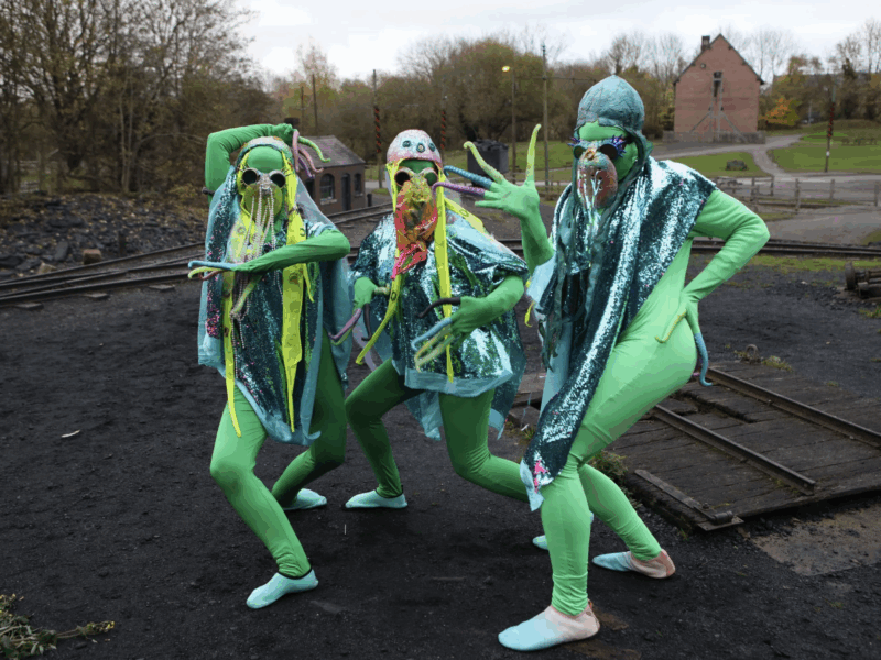 Three performers in bright green bodysuits and shiny blue capes pose dramatically outdoors near train tracks, wearing elaborate masks with tentacle-like decorations.