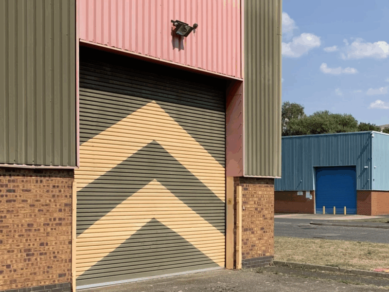 Industrial building with a large roller shutter door painted with yellow and green chevron stripes under a pink and olive green facade.