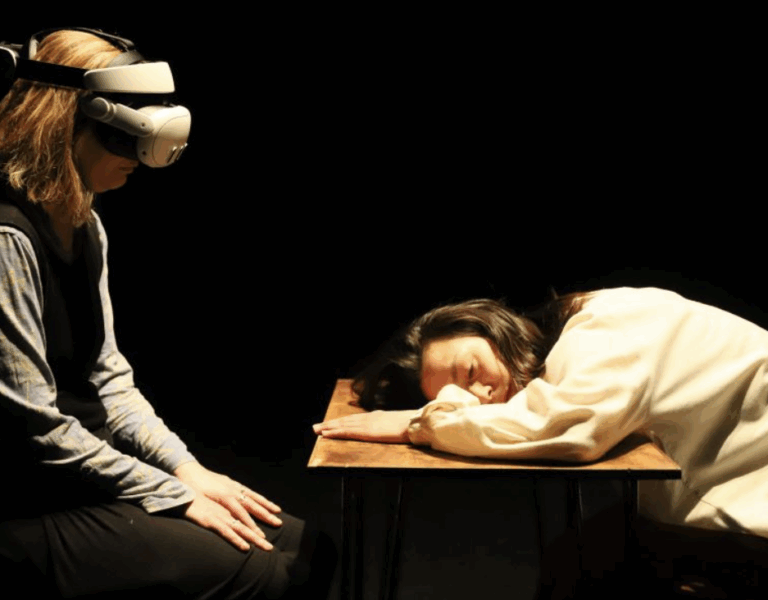 Person wearing a VR headset sits facing another person resting their head on a wooden table in a dimly lit room.
