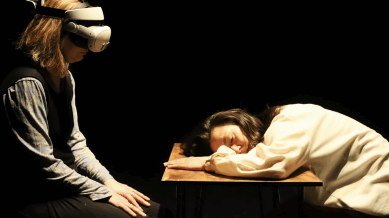 Person wearing a VR headset sits facing another person resting their head on a wooden table in a dimly lit room.