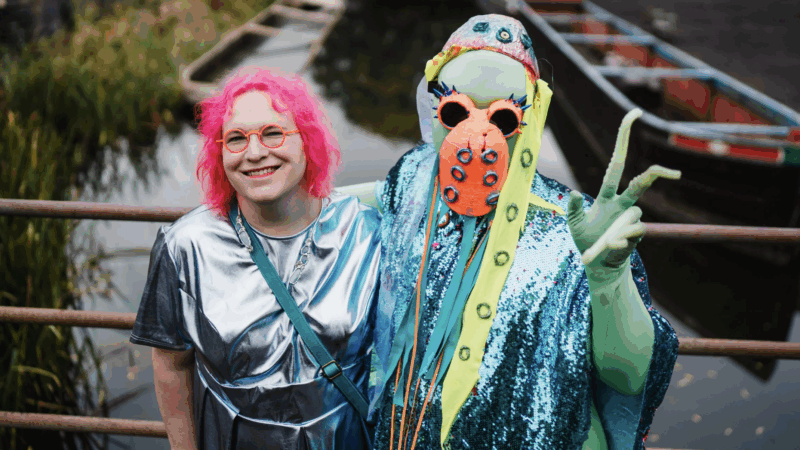 Two people in colorful costumes pose by a canal with boats, one wearing a shiny silver outfit and bright pink hair, the other in a blue sequined octopus-themed costume.