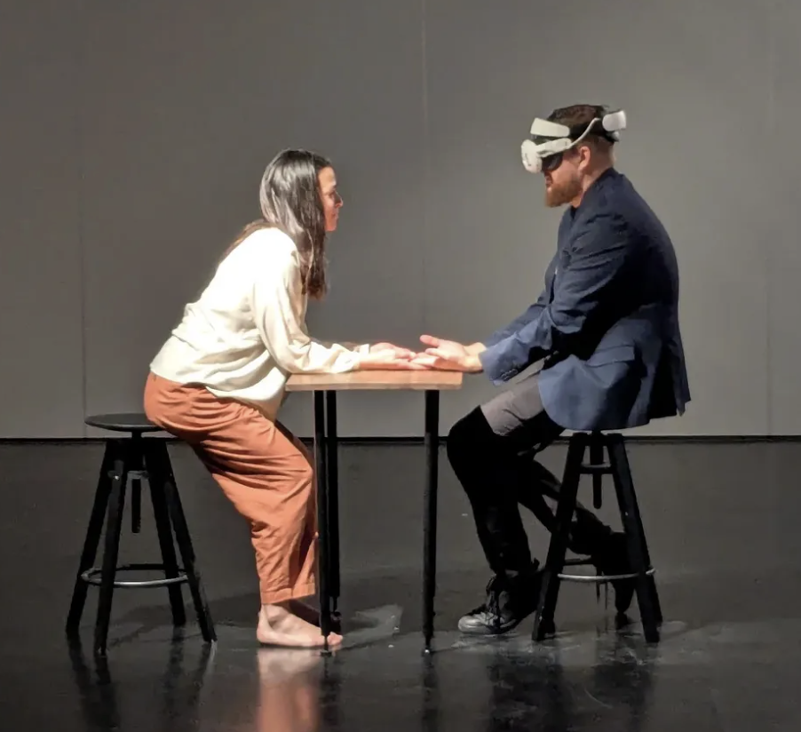 Two people sit at a table, across from each other. One wears a VR headset. Their arms are positioned on the table, barely touching hands.