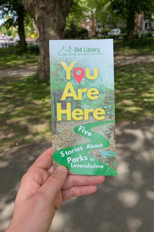 Photo of a hand holding a leaflet about 'You Are Here' in the foreground with a green space in the background.