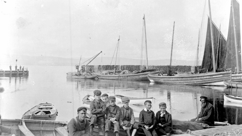 A black and white image of a group of boys sitting on a dock near small rowboats with sailboats and calm water in the background.
