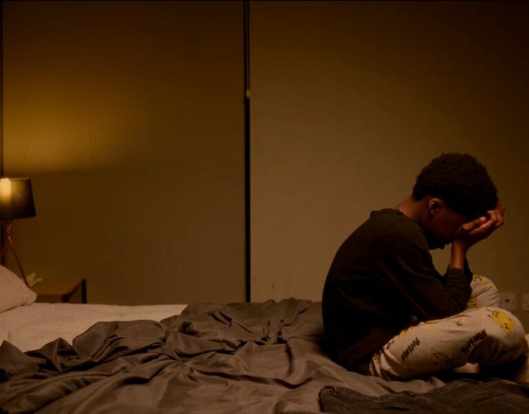 Still of 11 year old Malachi from 'Jury' sitting cross-legged on the edge of a bed, with his head in his hands.