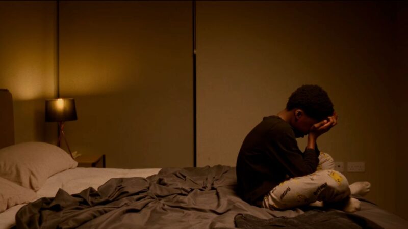 Still of 11 year old Malachi from 'Jury' sitting cross-legged on the edge of a bed, with his head in his hands.