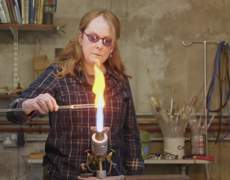 Woman in safety googles and a plaid shirt firing a piece of glass on an open flame.