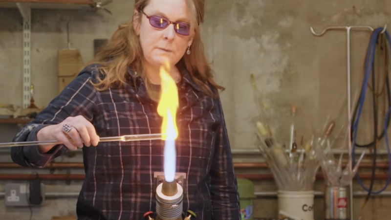 Woman in safety googles and a plaid shirt firing a piece of glass on an open flame.