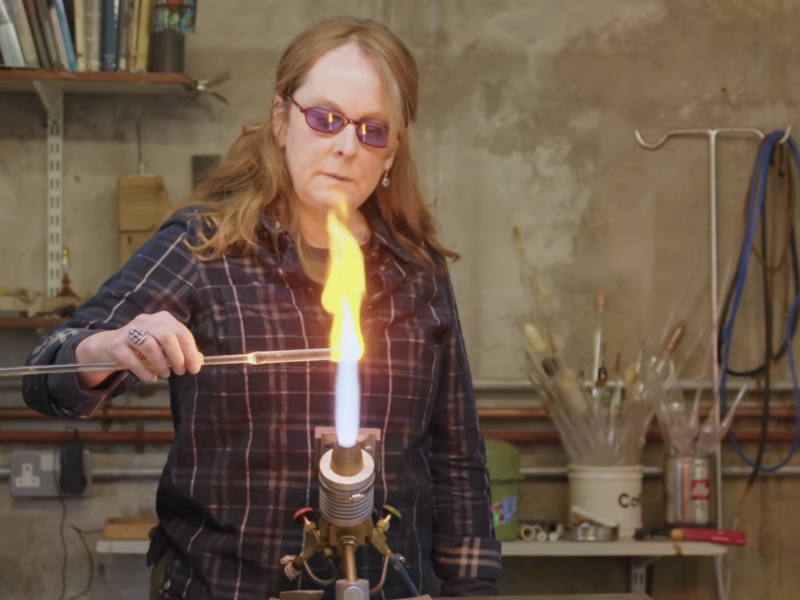 Woman in safety googles and a plaid shirt firing a piece of glass on an open flame.